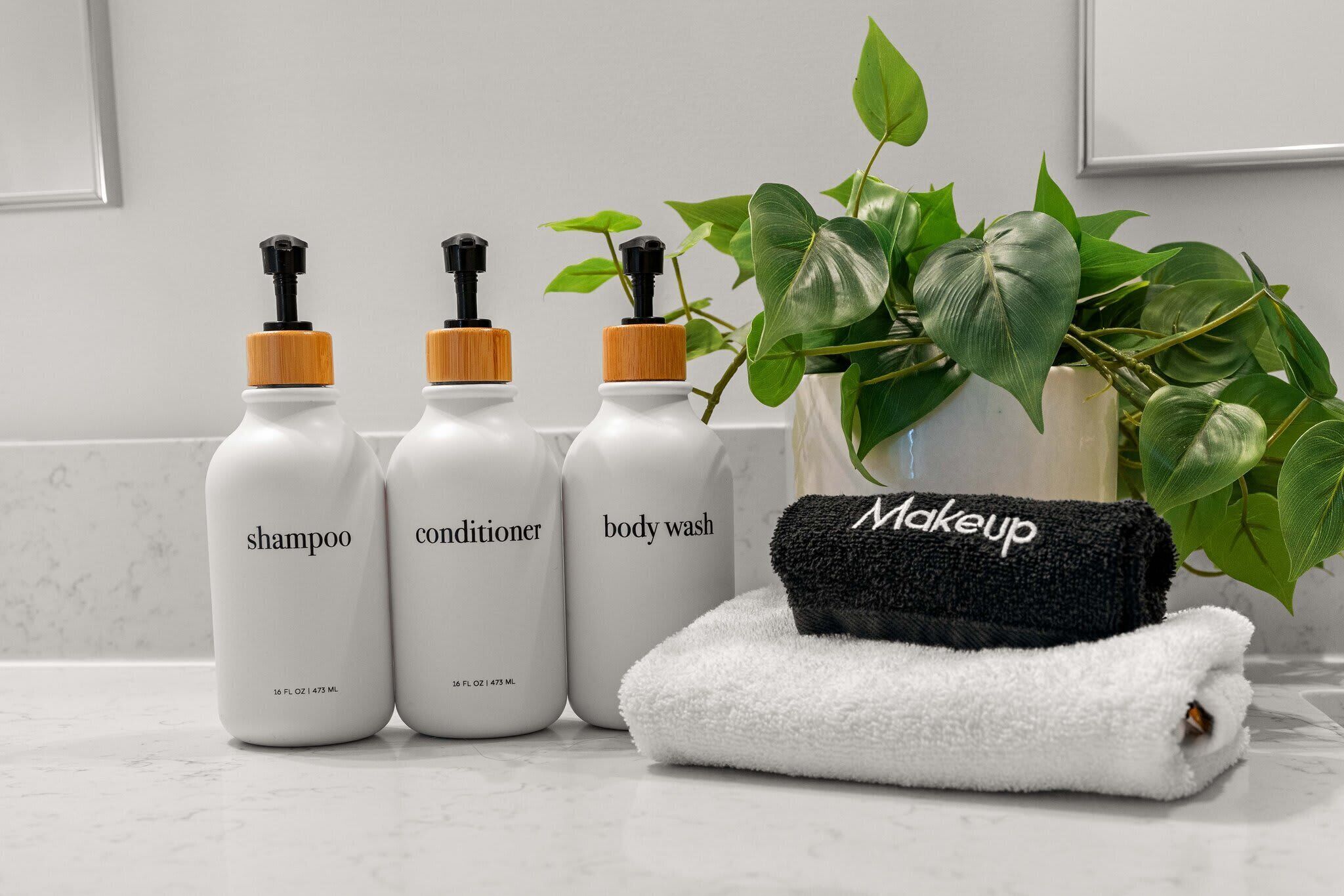Three bottles of shampoo, conditioner, and body wash, a black towel with 'Makeup' written on it, and a potted plant on a bathroom counter.