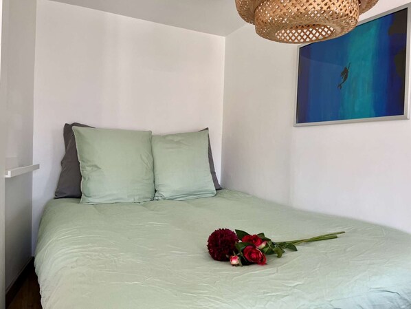 A bed with a light green bedspread and two pillows, a wicker lamp, and a painting of a diver on a blue background.