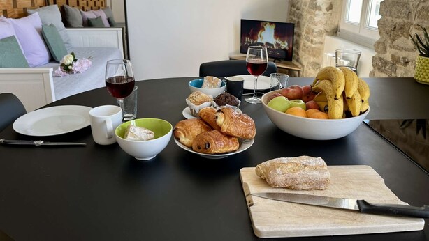 Ett matbord dukat med croissanter, bröd, frukt och vinglas i ett rum med stenmurar och en mysig sittgrupp.