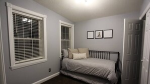 Un dormitorio con una cama, dos ventanas con persianas, una puerta y tres cuadros en la pared.