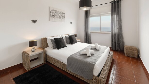 Quarto moderno com cama grande, mesa de cabeceira com abajur, vista para o mar pela janela e um detalhe decorativo na parede.