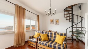 Sala de estar com sofá, lustre, escada em caracol e vista pela janela.