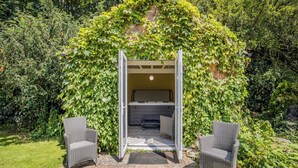 Un jacuzzi con cerramiento de vidrio rodeado de abundante vegetación, dos sillas de mimbre y un patio de piedra.