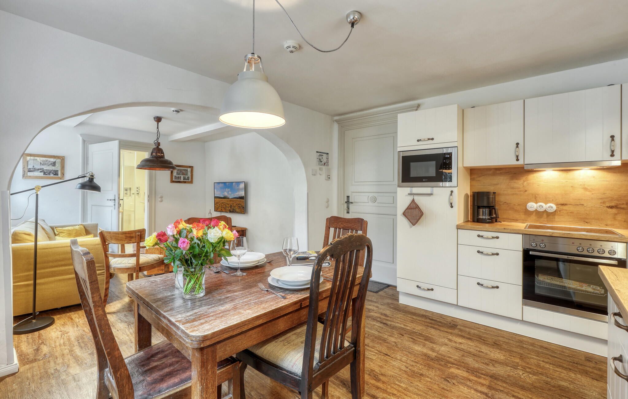 Zona pranzo con tavolo in legno, sedie e un vaso di fiori. Si vede una cucina con mobili e elettrodomestici bianchi.