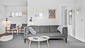 Ein modernes Wohnzimmer mit einem grauen Sofa, einem runden Couchtisch und einer weißen Stehlampe. An der Wand hängen zwei gerahmte Kunstwerke, und ein Fenster bietet Blick auf Bäume.