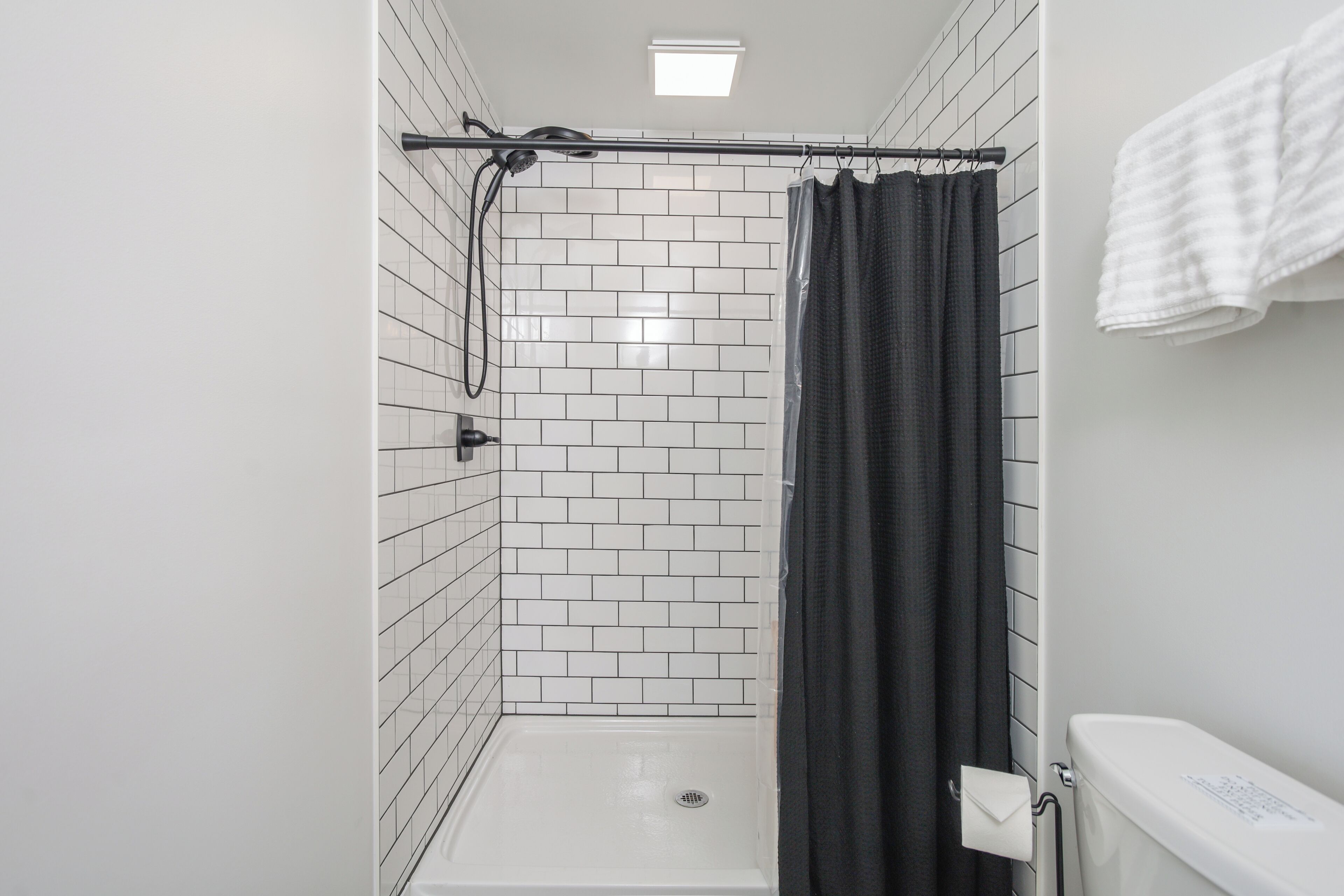 Zona de ducha con pared de azulejos blancos, ducha y cortina de baño negra.