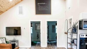A spacious room with a high ceiling, wooden beams, a mounted TV, a sofa, and a kitchen area with a stove. - Paddlewhack Cabin 4-The Gunwhale (Jefferson)