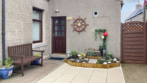 Un edificio de piedra con puerta de madera, un banco y un pequeño jardín con plantas y una estatua.