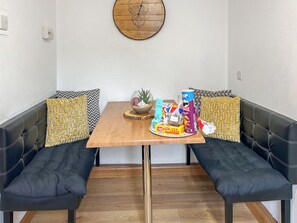 A dining area with a wooden table, two black cushioned benches, patterned cushions, a wall clock, and various food items on the table.