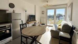 Eine moderne Küche mit Kühlschrank, Backofen und Esstisch. Im Wohnbereich befindet sich eine Couch und ein Sessel, mit Blick durch große Fenster.