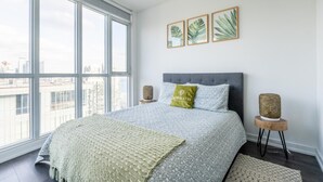 A modern bedroom with a large window, a grey upholstered headboard, a patterned bedspread, and a green pillow with text.