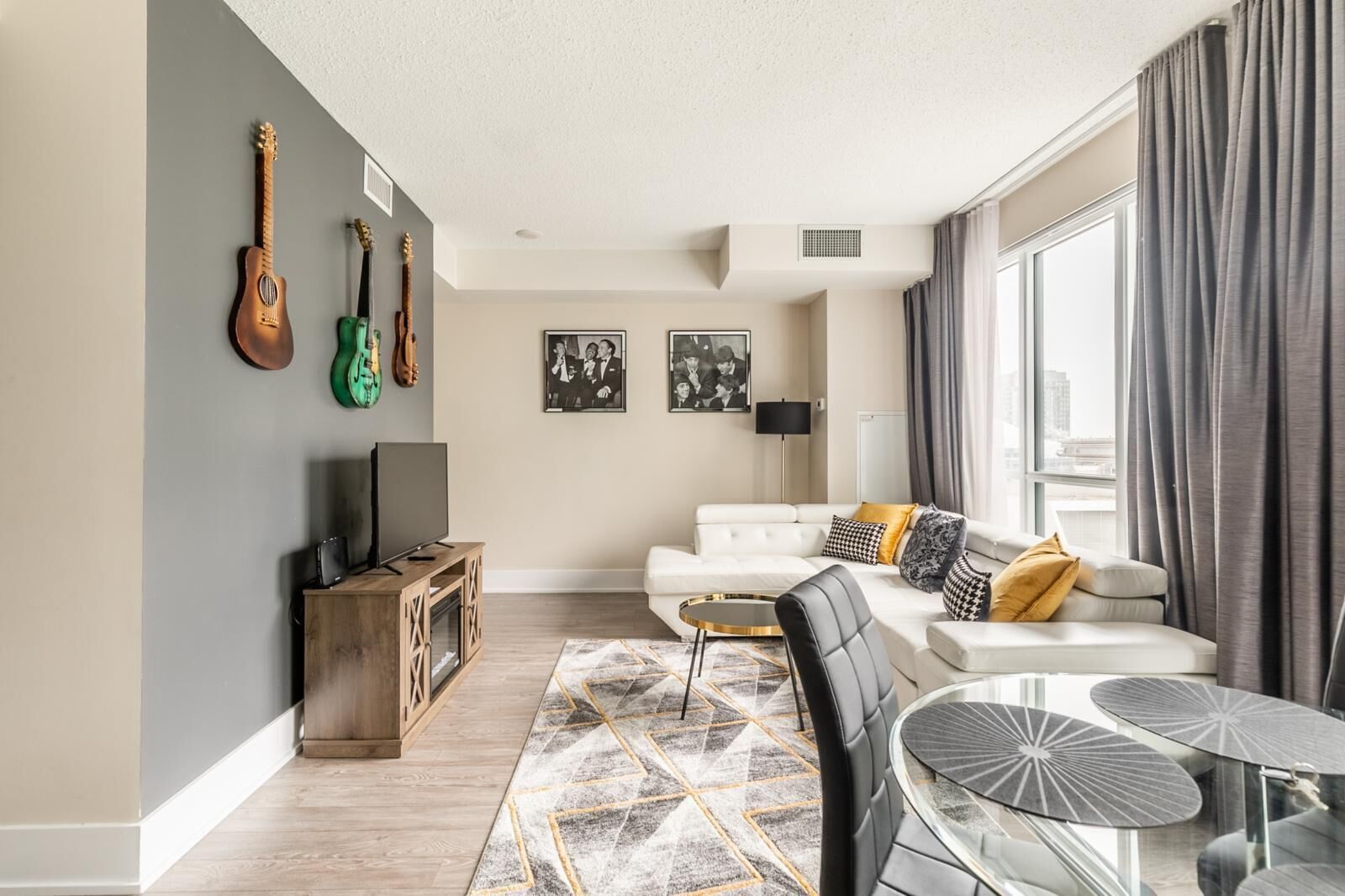 A modern living room with a white sofa, a glass coffee table, and a TV on a wooden cabinet. There are guitars on the wall and a city view through the window.