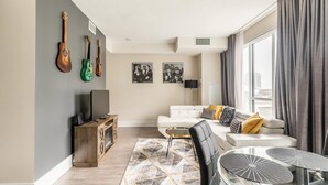 A modern living room with a white sofa, a glass coffee table, and a TV on a wooden cabinet. There are guitars on the wall and a city view through the window. - GLOBALSTAY. Stylish 1 Bedroom in Downtown Toronto (Toronto/Canada)