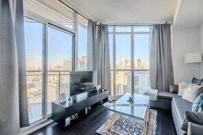 Ein modernes Wohnzimmer mit einem großen Fenster, einem Flachbildfernseher, einer schwarzen Unterhaltungseinheit, einem Glastisch und einem grauen Sofa sowie einem gemusterten Teppich.