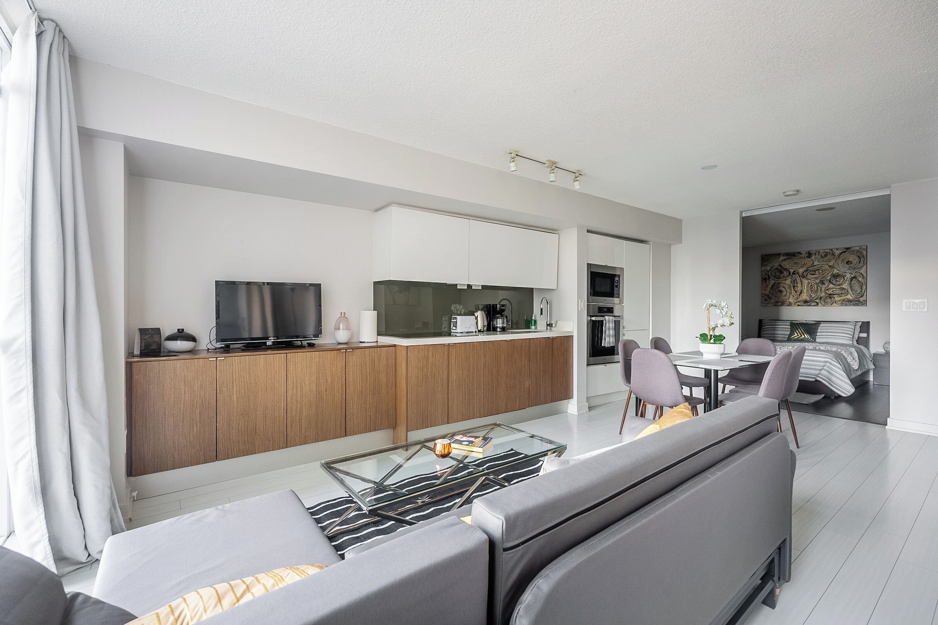 Een moderne woonkamer met een bank, een glazen salontafel, een televisie en een keuken met kasten en apparatuur.