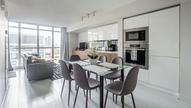 Une cuisine moderne avec des meubles blancs, un coin repas doté d’une table en verre et une vue sur la ville grâce à de grandes fenêtres.