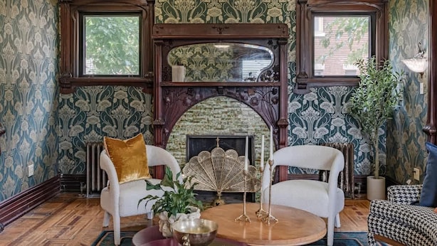 Une pièce avec un papier peint à motifs, une cheminée, deux fauteuils blancs, une table ronde avec un saladier et une plante en pot.