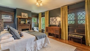Une chambre avec un grand lit, une commode en bois, un banc et un bureau. Il y a des fenêtres avec des rideaux et un tableau au mur.