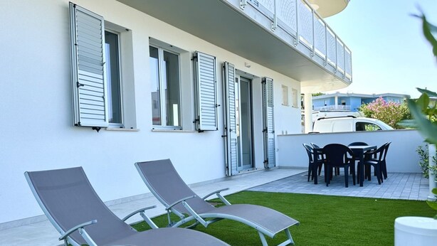 Un edificio moderno con un patio que tiene dos sillones, una mesa de comedor con sillas y una camioneta blanca estacionada cerca.