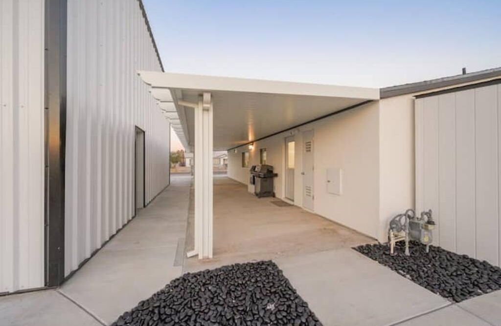 A covered outdoor area with a concrete pathway, metal buildings, and a small seating area with decorative items.