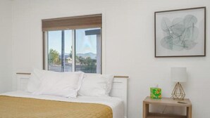 Une chambre à coucher avec un lit, une fenêtre donnant sur un beau paysage, une table de chevet avec une lampe et une œuvre d’art encadrée accrochée au mur.