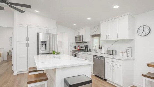 Une cuisine moderne avec des meubles blancs, un îlot central, des appareils en acier inoxydable et une horloge au mur.