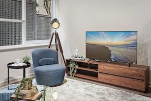 Un salon moderne avec une grande télévision, un fauteuil bleu, un meuble TV en bois, une lampe sur pied et une petite table avec une plante.