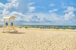 A beach with a lifeguard tower labeled '9 9', umbrellas, and people. - Gulf Shores Condo w/ Pool: Walk to Beaches! (Gulf Shores)