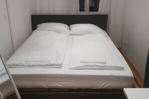 A neatly made bed with white linens and pillows, a wooden headboard, and a window with curtains.