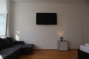 A room with a flat-screen TV mounted on the wall, a dark-colored sofa, a small bedside table with a lamp, and a wooden floor.