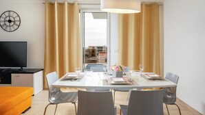 Un coin repas avec une table blanche dressée pour quatre personnes, un lustre moderne et une vue sur un quartier résidentiel grâce à de grandes fenêtres.
