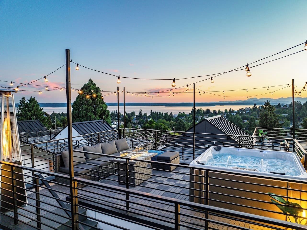 Een dakterras met een jacuzzi, buitenmeubels en lichtjes, tegen een zonsondergang.