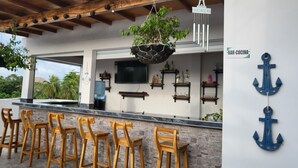 Un área de bar al aire libre con taburetes de madera, un macetero colgante y un televisor montado.