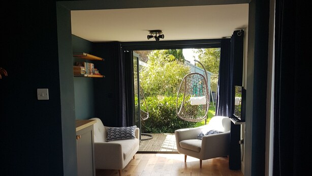 Une pièce comprenant un lit, deux canapés, une petite table et un fauteuil suspendu. Une porte donne accès à un espace extérieur agrémenté de verdure.
