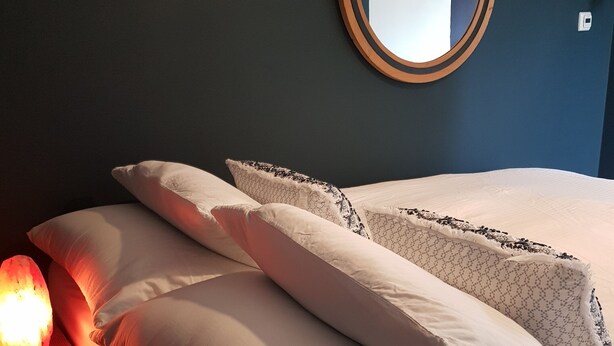 Una cama bien tendida con sábanas blancas y almohadas estampadas, un espejo redondo en la pared y una fuente de luz de tono rojizo en el suelo.