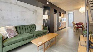 Un espacio de estar moderno con un sofá verde, una mesa de centro de madera y una zona de cocina con refrigerador y armarios.