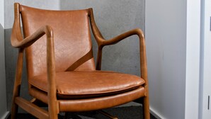 Una silla de cuero marrón con reposabrazos y patas de madera, ubicada frente a una pared gris.