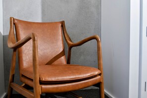 Un fauteuil en cuir marron, doté d’accoudoirs et de pieds en bois, placé contre un mur gris.