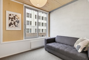 Ein modernes Wohnzimmer mit einem grauen Sofa, einem großen Fenster mit Metallstreben und einem Bauhaus-Poster an der Wand.