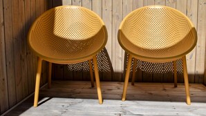 Dos sillas amarillas perforadas con patas de madera, ubicadas frente a una pared de madera con un patrón de sombras verticales.