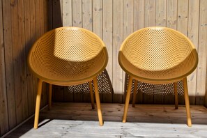 Deux chaises jaunes perforées, dotées de pieds en bois, sont placées contre un mur en bois présentant un motif d’ombres créé par des lattes.