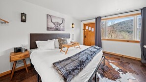 Ein Schlafzimmer mit einem großen Bett, einem Holfnachtkästchen, einem Nachttisch, einer Wandleuchte, einem gerahmten Bild, einem Fenster mit Vorhängen und Blick auf eine gebirgige Landschaft.
