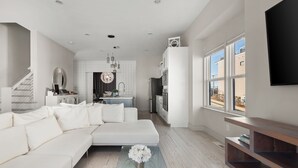 Un salon moderne avec un canapé d’angle blanc, une table basse en verre ornée de fleurs blanches et un coin cuisine doté de meubles blancs et d’une grande fenêtre.