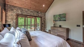 Une chambre avec un mur en pierre, un grand lit, une commode en bois et un téléviseur fixé au mur.