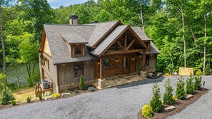 A log cabin with a stone foundation, wooden exterior, and a gravel driveway. - Rushing River Retreat - Waterfront on Toccoa | Hot Tub | Game Room | Modern Luxury (Mineral Bluff)