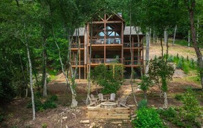 Une cabane en bois à plusieurs niveaux, entourée d’arbres, avec une terrasse et un espace salon extérieur.