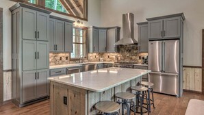 A modern kitchen with grey cabinets, a large island with stools, a stainless steel refrigerator, and a stone backsplash.