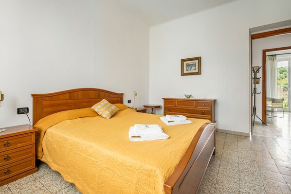 A neatly made bed with a yellow bedspread and two folded towels, a wooden nightstand with a lamp, a small table with a picture frame, and a coat rack with a mirror.
