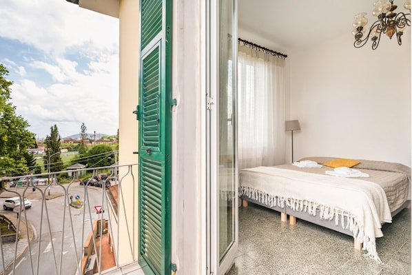 A balcony with a view of a street and trees, a bed with a white bedspread and a yellow pillow, and a chandelier.