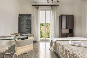 A hotel room with a bed, a glass table with chairs, a dark cabinet, and a view of greenery through the window.